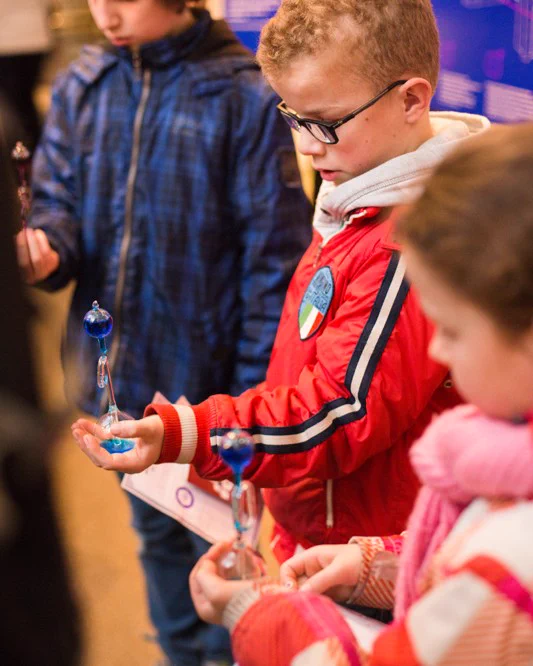 Three children stand diagonally to the camera holding hand boilers with blue liquid bubbling into the top chamber.