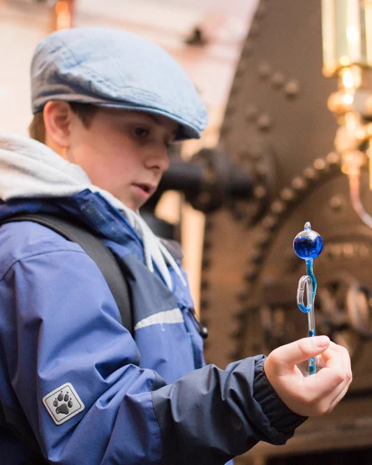 A child in a blue jacket and flat cap holds a hand boiler with blue liquid bubbling into the top chamber.