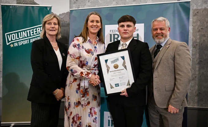 A photograph of four people dressed in smart clothes. In the middle is woman in a floral dress shaking hands with a man in a black suit holding a framed certificate