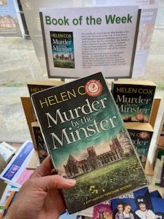 Image is a book held by a hand. The cover depicts a view across a field to a large church.
