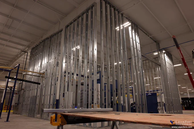 The image shows large steel beams going from floor to ceiling around a blue Powder Coating area. There are gaps in between the beams as the wall is still being built. There's a large gap to the right of the image where a door will go. In the foreground of the image there is a table to measure material. The white LayerZero Power Systems Inc., logo is in the right corner as well as white text with the LayerZero website address, www.LayerZero.com