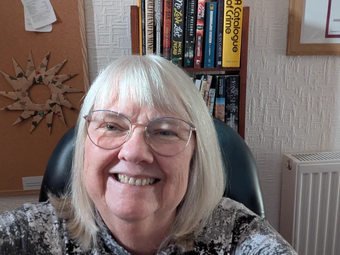 Maura has short white hair and is wearing glasses. She's smiling and looking into the camera, there is a bookcase behind her with a number of different books on it.