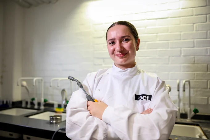 A female student wearing a white lab coat which has an embroidered UCL logo and holding lab equipment. 