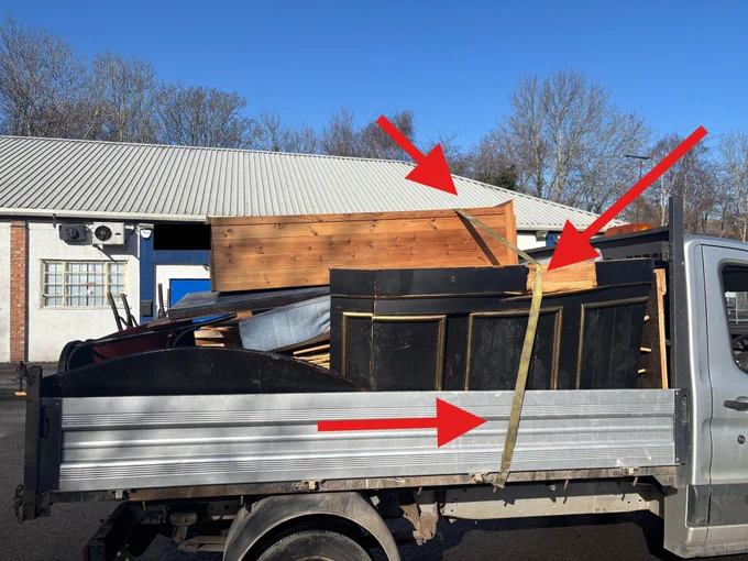Side view of a flatbed truck, overloaded wth household goods, being held with a single strap.