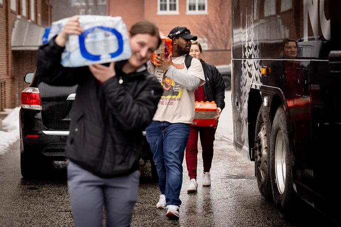 A line of people walk towards a bus on right. Person in foreground is carrying a small pallet of water bottles.