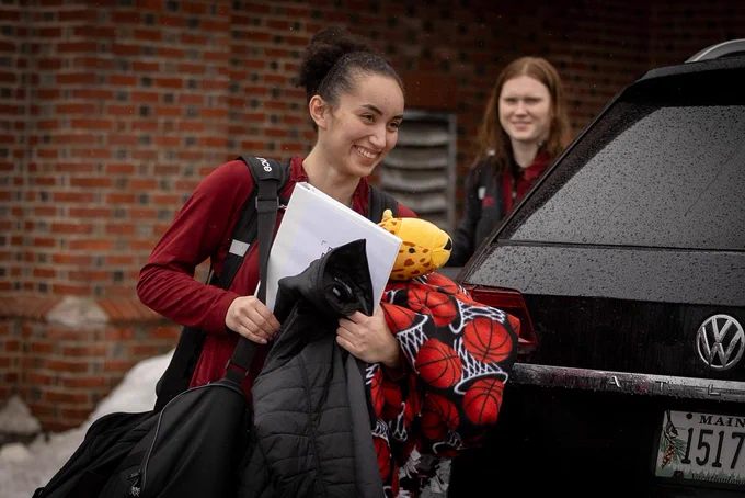 A student smiles while holding her jacket and gear as she makes her way onto the bus as the Bates women's basketball team head to Scranton, Pa., for the NCAA DIII Tournament.