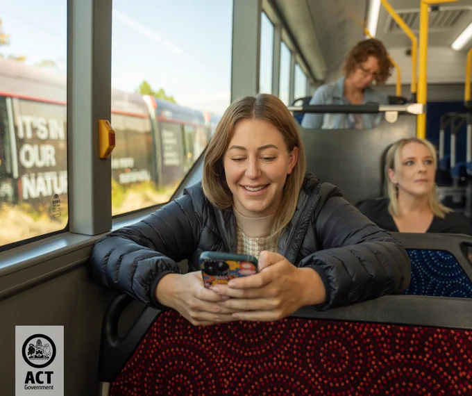 Person smiling while looking at a smartphone, sitting on a bus, with another individual in the background.