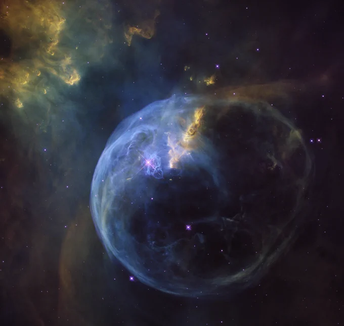 A Hubble Space Telescope image of the Bubble Nebula, showcasing a large, spherical shell of glowing gas with a distinct blue hue. Wisps of yellow and gold are visible along the edges and within the bubble's structure.