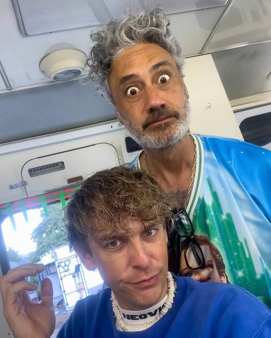 Taika Waititi in a Wizard of Oz shirt and silver chain stands with his eyes wide, reading glasses hanging from his shirt, and his grey and white hair standing on end. The man sitting in front of him has curly hair a blue shirt and they are both looking at the camera. 