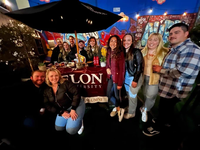 A group of alumni from San Francisco gathered in front of an Elon University Alumni banner, smiling for a photo at a casual outdoor event.