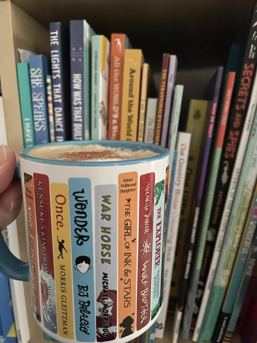 A coffee mug which is decorated with the spines of famous children’s books being held in front of a shelf of kids books.