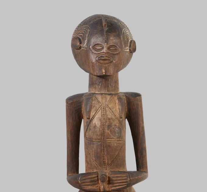 A carved wooden figure in the shape of a woman. The figure has a round stylised head with a pointed chin, and almond shaped eyes, ears and mouth. A series of arched lines are carved above each ear, representing her braided hair. The figure stands with her hands on either side of her navel, which protrudes outwards. A geometric pattern - possibly representing scarification - consisting on a diamond shape split into quarters by a cross is carved between her breasts, which are stylised as downward pointing triangles.