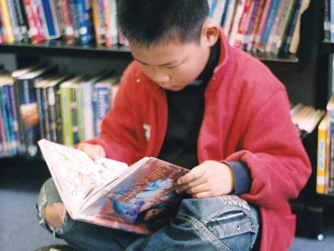 Photo of a young boy sat in front of bookshelves, reading a book.
