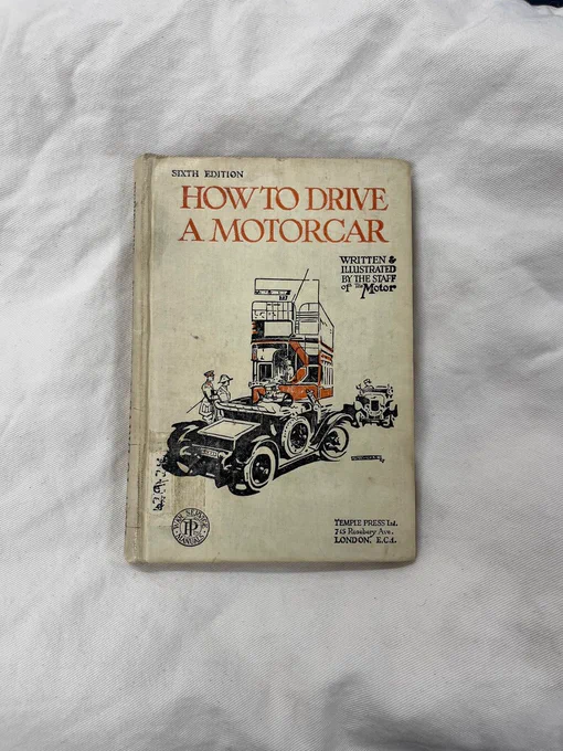 A colour photograph of a book titled 'How to Drive a Motorcar'. The cover depicts a motorcar turning right with a red bus in front of it and another motorcar coming down the opposite side of the road.