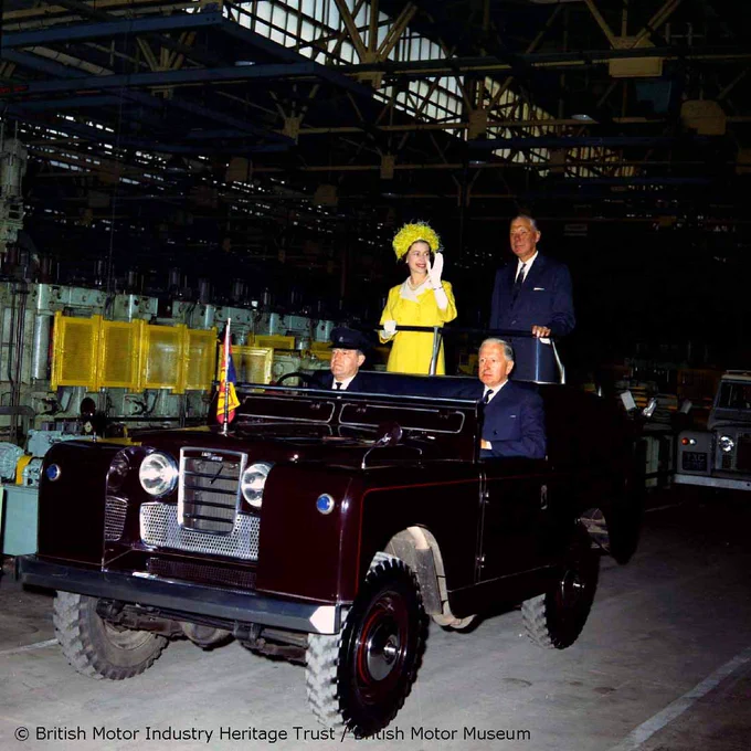 A colour photograph of Queen Elizabeth II wearing a yellow dress and hat and waving while standing in an open top ceremonial Land Rover in a factory. A man stands next to her and two more men sit in the seats at the front of the vehicle looking forward. 