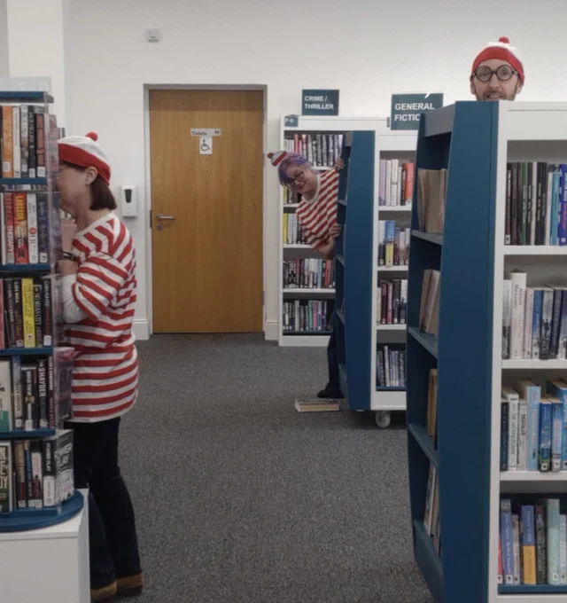 Three people dressed as Where’s Wally hiding behind bookshelves in the library.