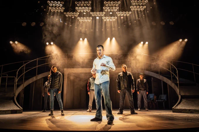A group of people in dark clothes stand tall, with legs wide. They look out, defiant. Behind them sits a stage set with a curving bridge and pairs of spotlights shine out.