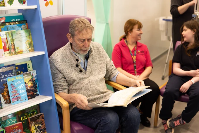 Michael Rosen reading his book, behind him is an Evelina London play specialist and a patient