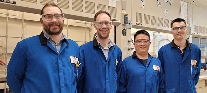 Four UTSA engineers smiling in a laboratory, wearing blue lab coats with UTSA logos.