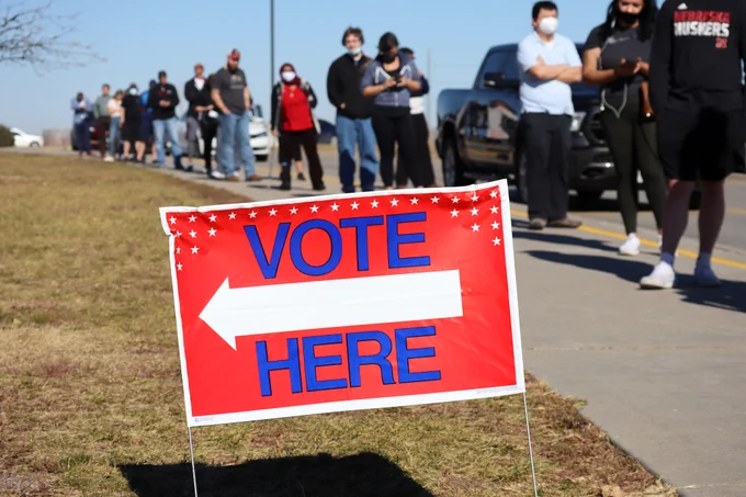 Nebraska voters wait in line to cast their ballots. In the front of the picture, a sign reads "VOTE HERE." 
