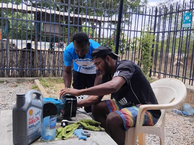Two individuals are working together on a mechanical project outdoors, near a metal fence. They are using tools and are focused on repairing a small engine placed on a wooden board. A bottle of Shell Helix HX7 oil and a white chair are also visible in the scene.