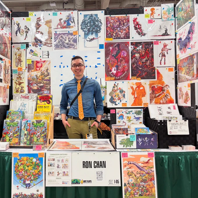 Photo of me behind my convention table displaying art prints and comics. I'm an Asian guy with glasses, wearing a denim shirt with a yellow tie.