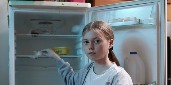 A young girl stands in front of an open refrigerator, reaching for an item inside.