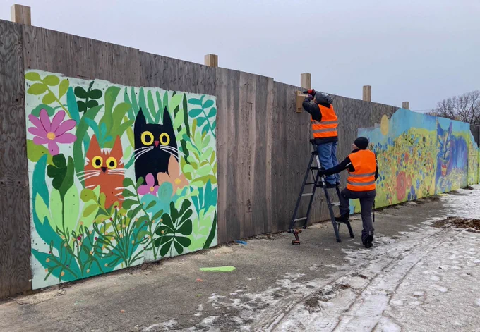 Two people in orange vests, one standing on a ladder, install a wooden bird house on a wooden fence surrounded by brightly coloured murals.