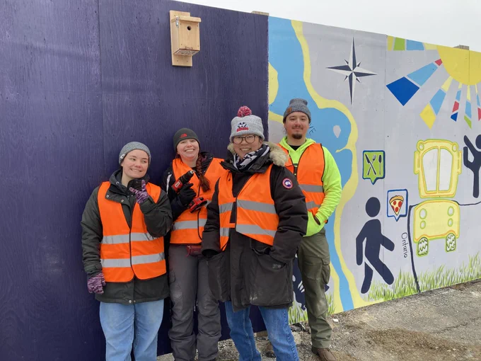 Two people in orange vests, one standing on a ladder, install a wooden bird house on a wooden fence surrounded by brightly coloured murals.