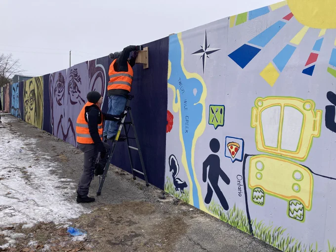 Two people in orange vests, one standing on a ladder, install a wooden bird house on a wooden fence surrounded by brightly coloured murals.