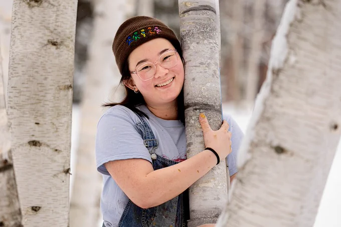 Sophie Hafter, wears a hat and smiles as she hugs her artificial birch tree amidst a group of real birch trees on Bates' Alumni Walk.