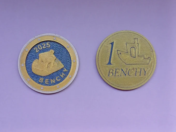 Two views (anverse and reverse) of a Benchy maker coin. Anverse is gold, blue and silver, and has a golden Benchy with the legend "2025 Benchy" on blue gyroid waves, surroinded by 10 stars on a golden ring.
The reverse features the legend "1 Benchy", with an outline of the Benchy side.