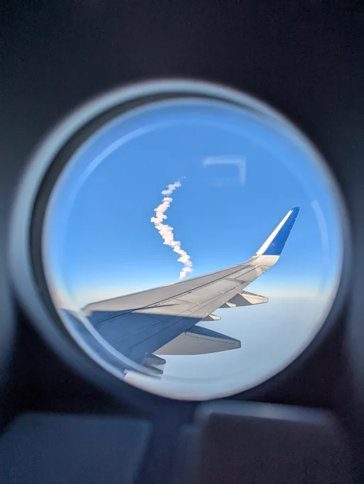A picture through a porthole window in an airplane door showing a rocket plume alongside the airplane wing and winglet
