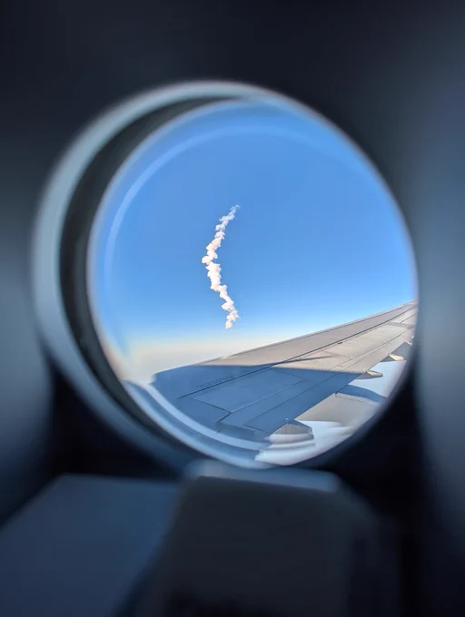 A porthole window in an airplane door, showing the airplane wing as well as a plume of rocket exhaust