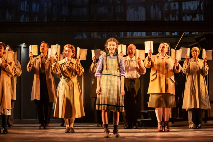 A young actress centre stage in front of a group of actors each holding up a book, all dressed in 1930s period costumes