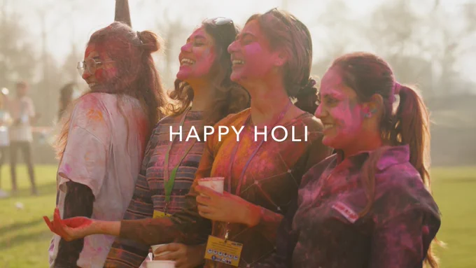 4 women smiling, covered in colourful Holi powder. Text reads "Happy Holi"