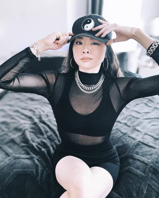 Ruibo Qian sits on the edge of a bed in a black transparent shirt with a black bra underneath. Her black hat has a ying/yang on it. She wears silver necklaces, earrings and bracelets. 