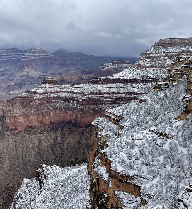 snow covering higher elevation cliffs and slopes within a vast, mile deep canyon landscape. Storm clouds fill the sky.  