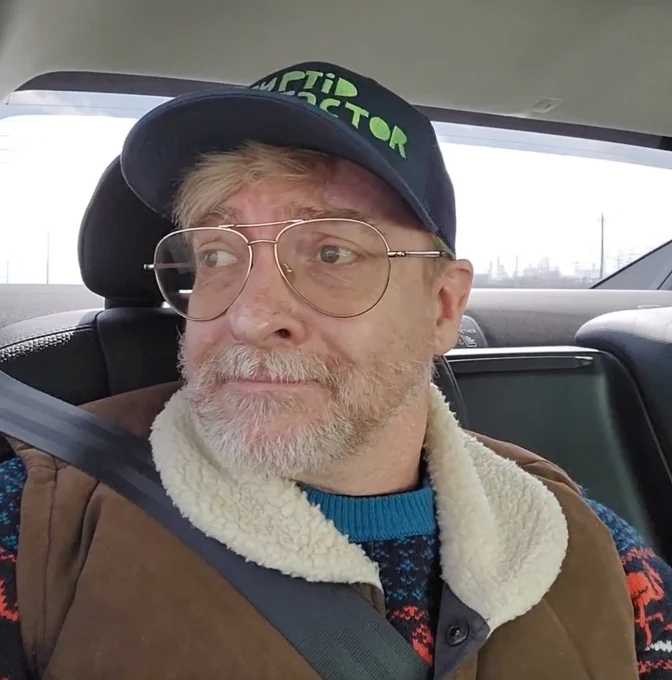 Rhys Darby sits in the back seat of a car in a flamingo sweater with a brown jacket on top. He wears a Cryptid Factor Hat. He wears glasses and looks out the window. 