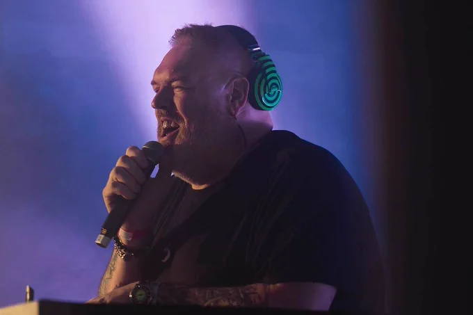 Kristian Nairn Stands in a hall in front of his DJ equipment, speaking into a microphone. 