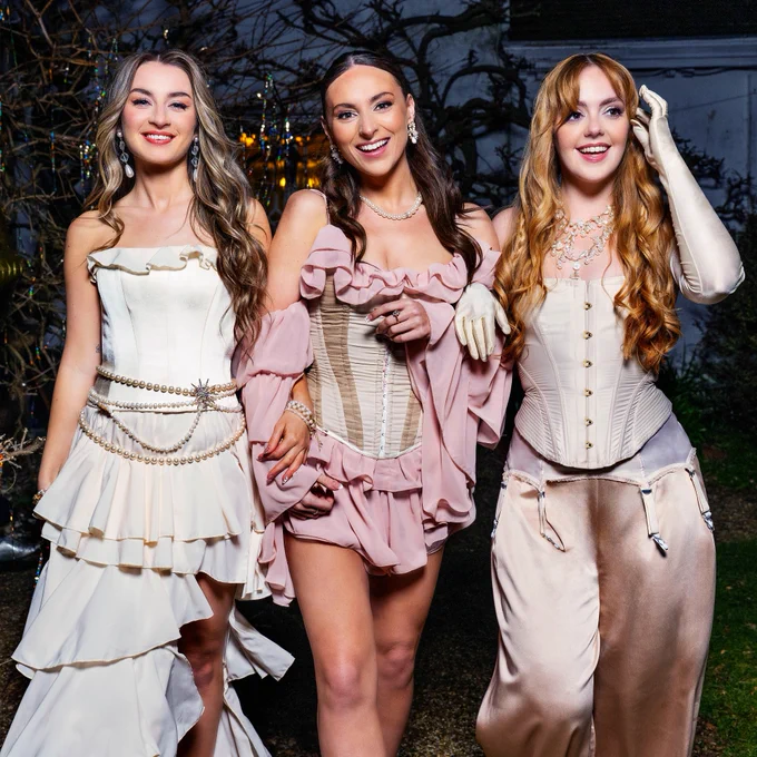 Three young women in light pastel colour corsets, dresses and trousers walk arm in arm, smiling for the camera.