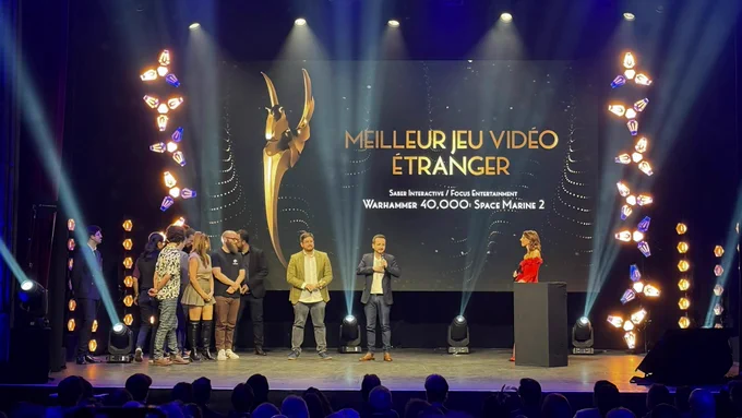 A photograph captures an awards ceremony on a stage with a large screen displaying "Meilleur Jeu Vidéo Étranger" (Best Foreign Video Game) awarded to Warhammer 40,000: Space Marine 2 by Saber Interactive and Focus Entertainment. A group of developers and representatives, stand on the stage. A woman in a red dress stands behind a podium. The stage is illuminated with spotlights and decorative lighting fixtures, while an audience watches in the foreground.