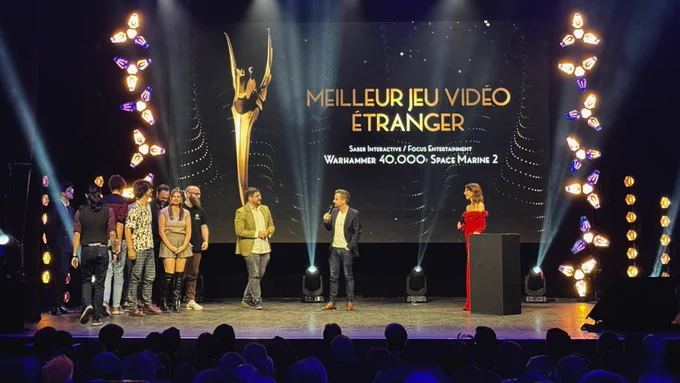 A photograph captures an awards ceremony on a stage with a large screen displaying "Meilleur Jeu Vidéo Étranger" (Best Foreign Video Game) awarded to Warhammer 40,000: Space Marine 2 by Saber Interactive and Focus Entertainment. A group of developers and representatives, stand on the stage. A woman in a red dress stands behind a podium. The stage is illuminated with spotlights and decorative lighting fixtures, while an audience watches in the foreground.