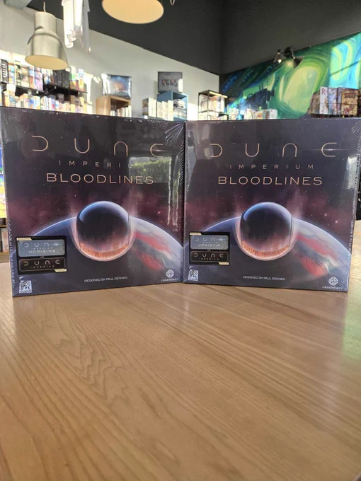 Two copies of the Dune Imperium Bloodlines board game on a table in the board game shop Ancient Robot Games. A robot mural and shelves of board games are visible in the background.
