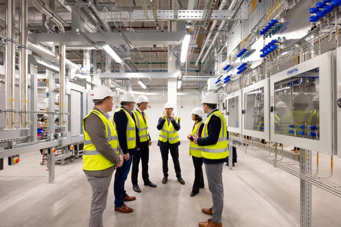 Secretary of State Peter Kyle and State for Wales Jo  Stevens among others in hard hats and high-visibility vests discussing in a modern industrial facility with complex machinery and piping.
