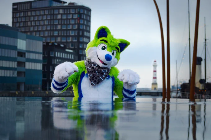 Green husky fursuiter standing behind a fountain which looks like a huge water bowl. In the background are office buildings and a light house.