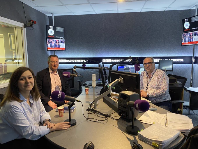 Three people sit at studio desks, microphones in front of them. Behind are signs reading "BBC Radio Bristol".