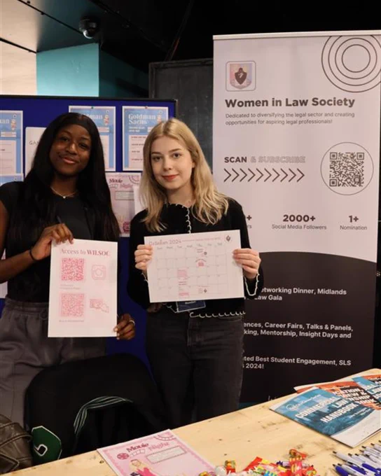 Image of two students smiling at the camera whilst holding up posters who are a part of the 'Women in Law Society'.