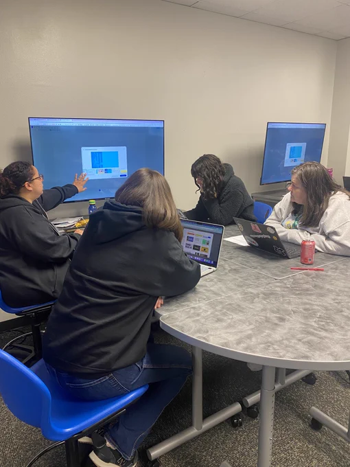 4 women sitting at a table while 1 presents on a Tv and the others are watching.