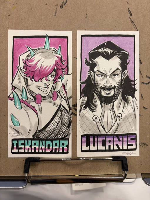 Photo of two small ink and marker sketches. One is a buff jock with pink hair and teal horns. The other is a man with a beard and long black hair in a vest.